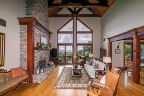 A spacious living room with a stone fireplace, wooden beams, and large windows offering a view of the outdoors. - Haven at Headwaters Long-range Mountain Views, Pet Friendly With Game Room (Elk Park)