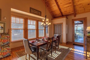 A dining area with a wooden table set for four, a chandelier, and a view through large windows. - B Rock Ridge at Echota - Townhome With Private hot tub (Banner Elk)