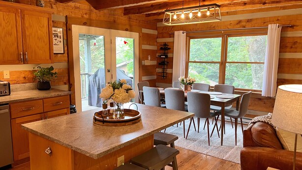 Una cocina tipo cabaña con armarios de madera, encimera de granito, zona de comedor con mesa y sillas, y un ventanal con cortinas.