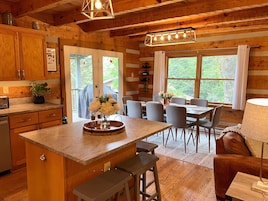 A cabin-style kitchen with wooden cabinets, a granite countertop, a dining area with a table and chairs, and a large window with curtains.