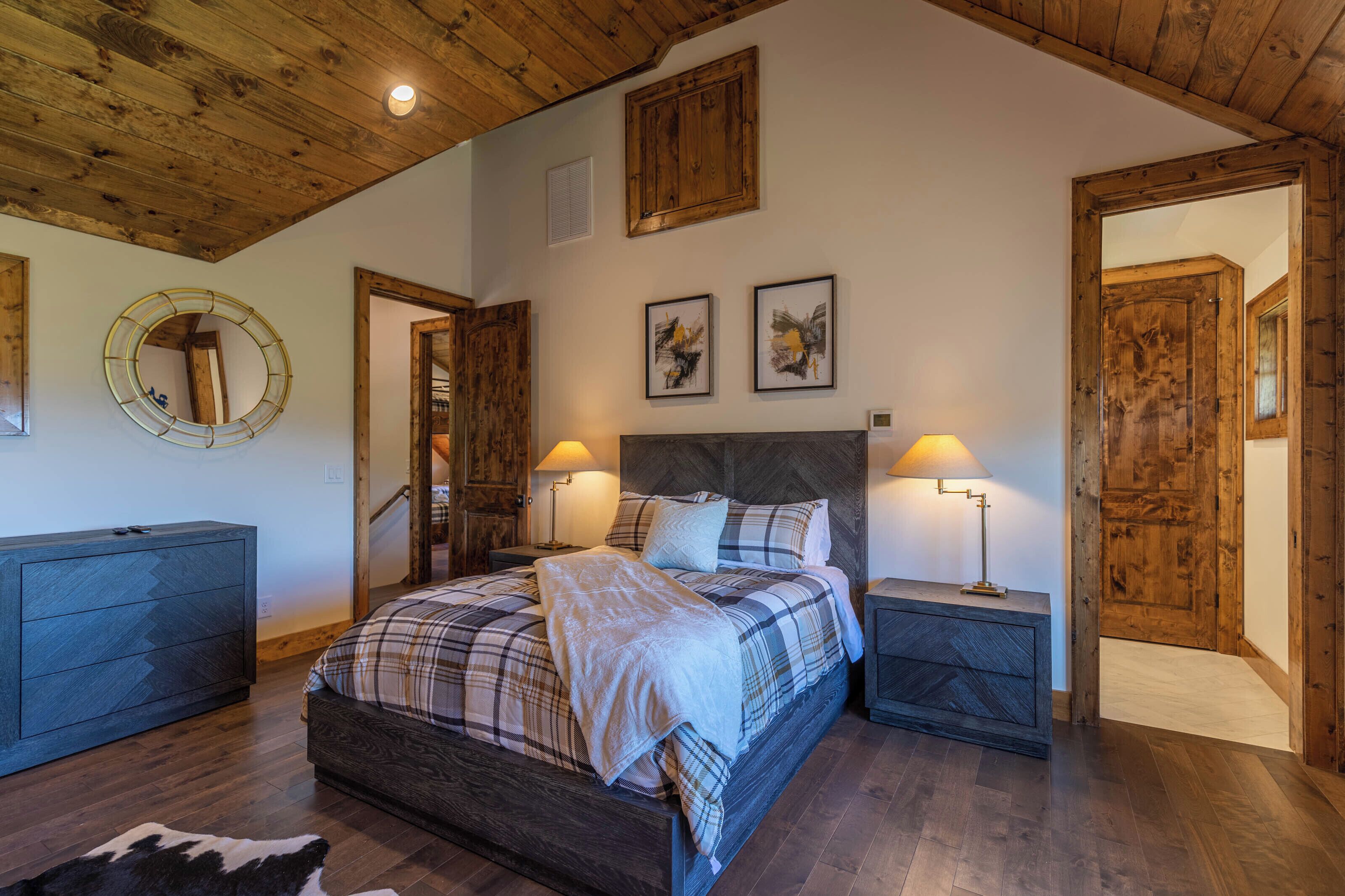 A bedroom with a wooden ceiling, a bed with plaid bedding, two bedside lamps, and a mirror on the wall.