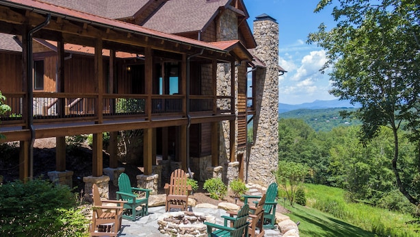 A large log cabin with a stone chimney, a covered porch with outdoor seating, and a scenic view of the surrounding forest. - Breezeway Lodge at Eagles Nest (Banner Elk)