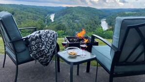 Een tafel met een haardvuur, twee glazen rode wijn en een schaal met hapjes, met uitzicht op een rivier en beboste heuvels.