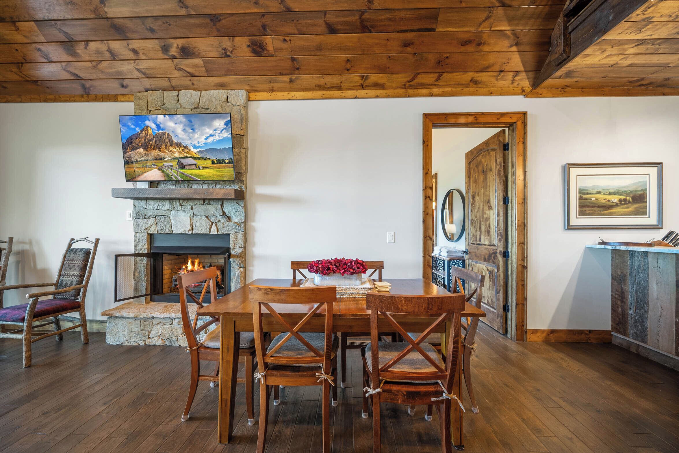 Zona pranzo con camino in pietra, soffitto in legno e un dipinto di un paesaggio montano.