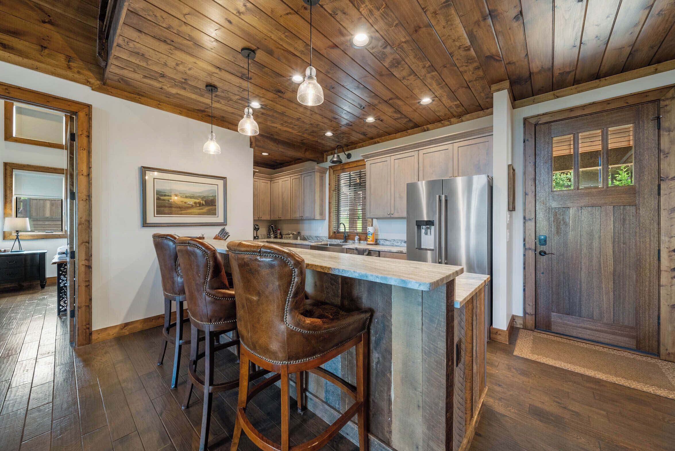 A kitchen with wooden ceiling, bar stools, stainless steel appliances, and a wooden door.