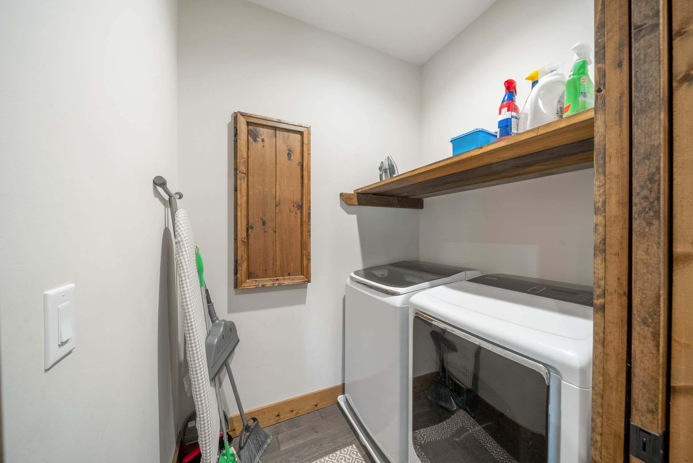A compact kitchen with a washing machine, a drying rack, and a wooden shelf with cleaning supplies.