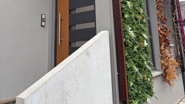 Un bâtiment moderne à la façade grise, doté d’une porte en bois et d’un jardin vertical agrémenté de plantes séchées.