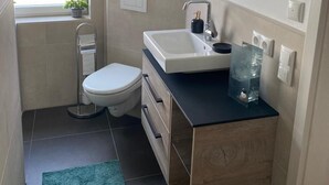 Une salle de bain moderne avec des toilettes, un lavabo et un miroir. Des plantes sont posées sur le rebord de la fenêtre et un tapis vert recouvre le sol.