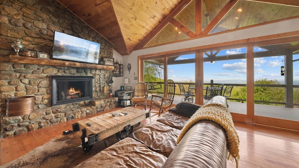 Un salón acogedor con chimenea de piedra, techo de madera y un amplio ventanal con vistas panorámicas.