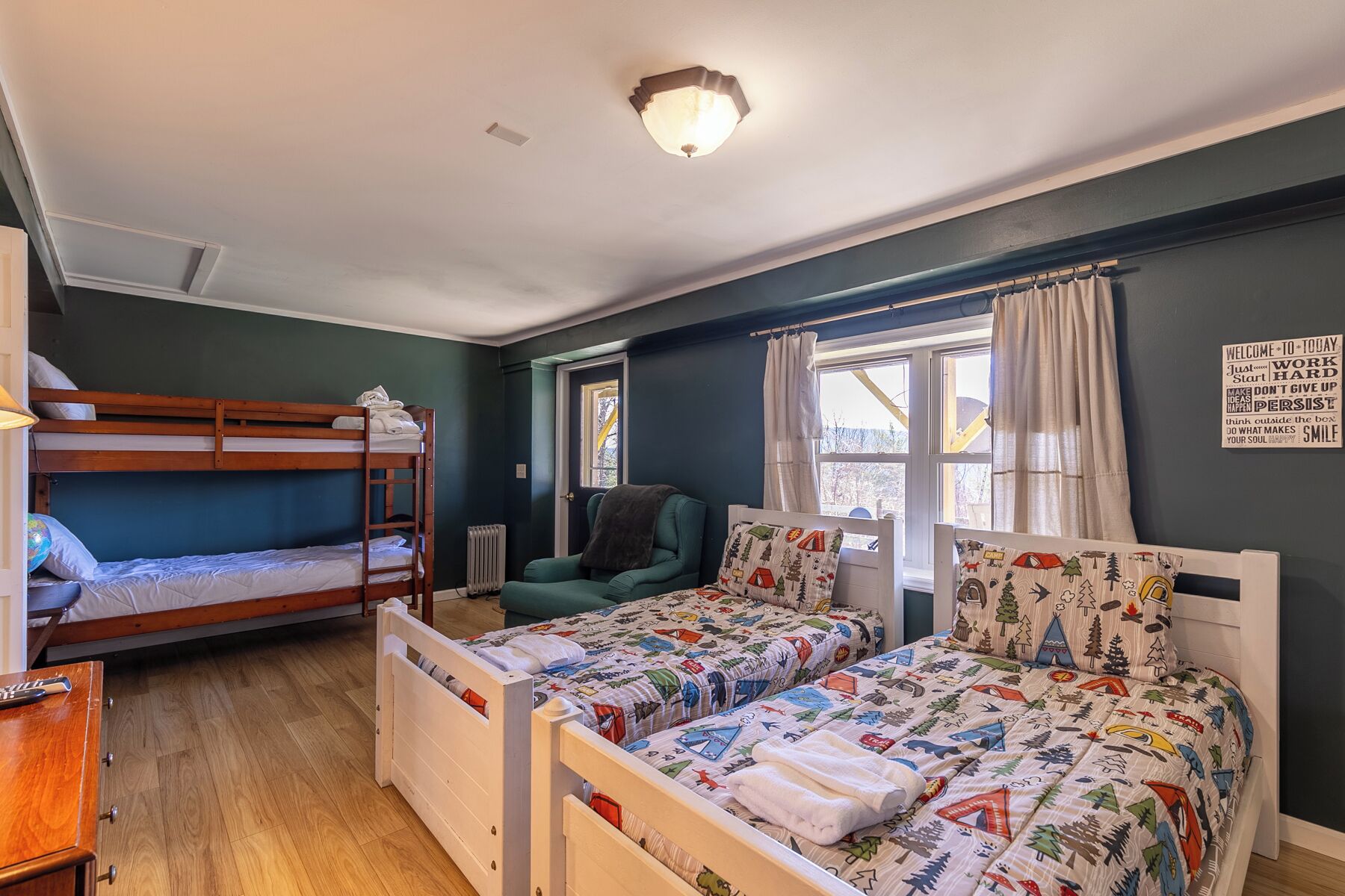 A room with bunk beds, a green armchair, a bed with a forest-themed duvet, and a motivational quote wall.