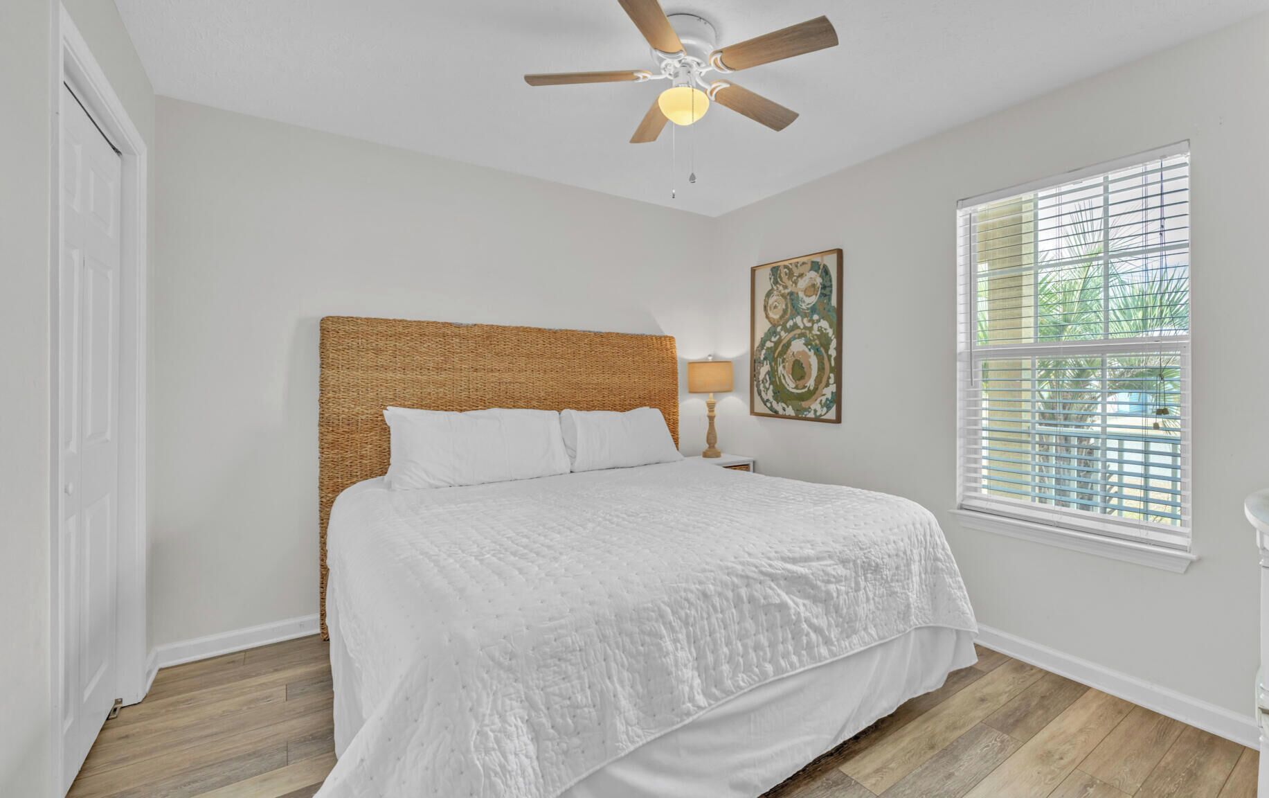 A bedroom with a bed, a ceiling fan, a window with blinds, and a wall-mounted lamp.