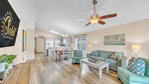 A spacious living room with a ceiling fan, a dining area with a kitchen in the background, and a large window with blinds. - Cottage on Cabana Private Gated Neighborhood (Panama City Beach)