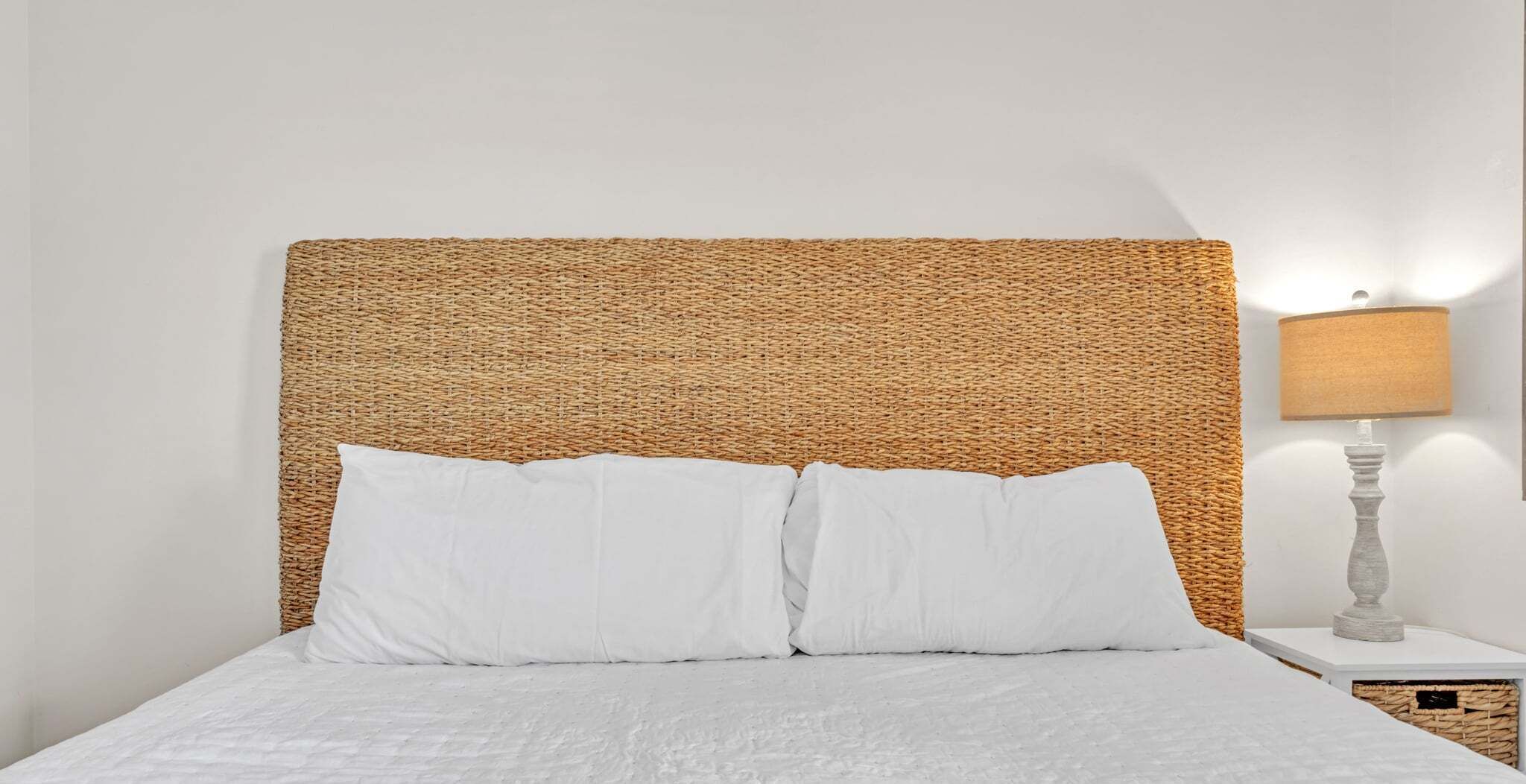 A bed with white bedding, a woven headboard, and a bedside lamp on a white nightstand.