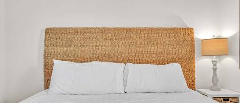 A bed with white bedding, a woven headboard, and a bedside lamp on a white nightstand.