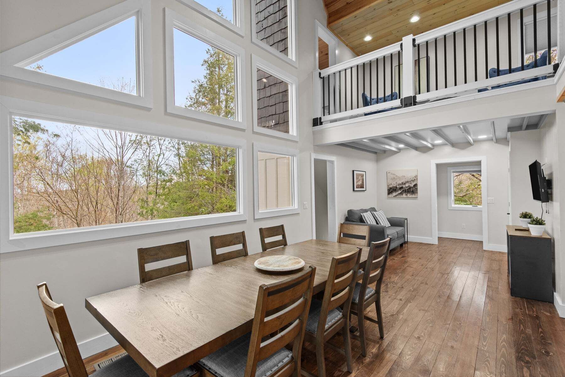 A spacious dining area with a wooden table and chairs, large windows, a balcony with a railing, and a cozy seating area with a sofa and cushions.