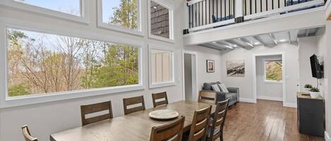 A spacious dining area with a wooden table and chairs, large windows, a balcony with a railing, and a cozy seating area with a sofa and cushions.
