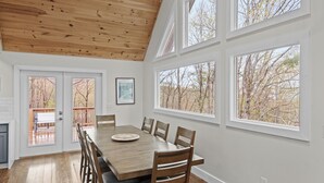 A dining room with a wooden table and chairs, large windows with a view of trees, and a white ceiling. - Elevation Escape (Banner Elk)
