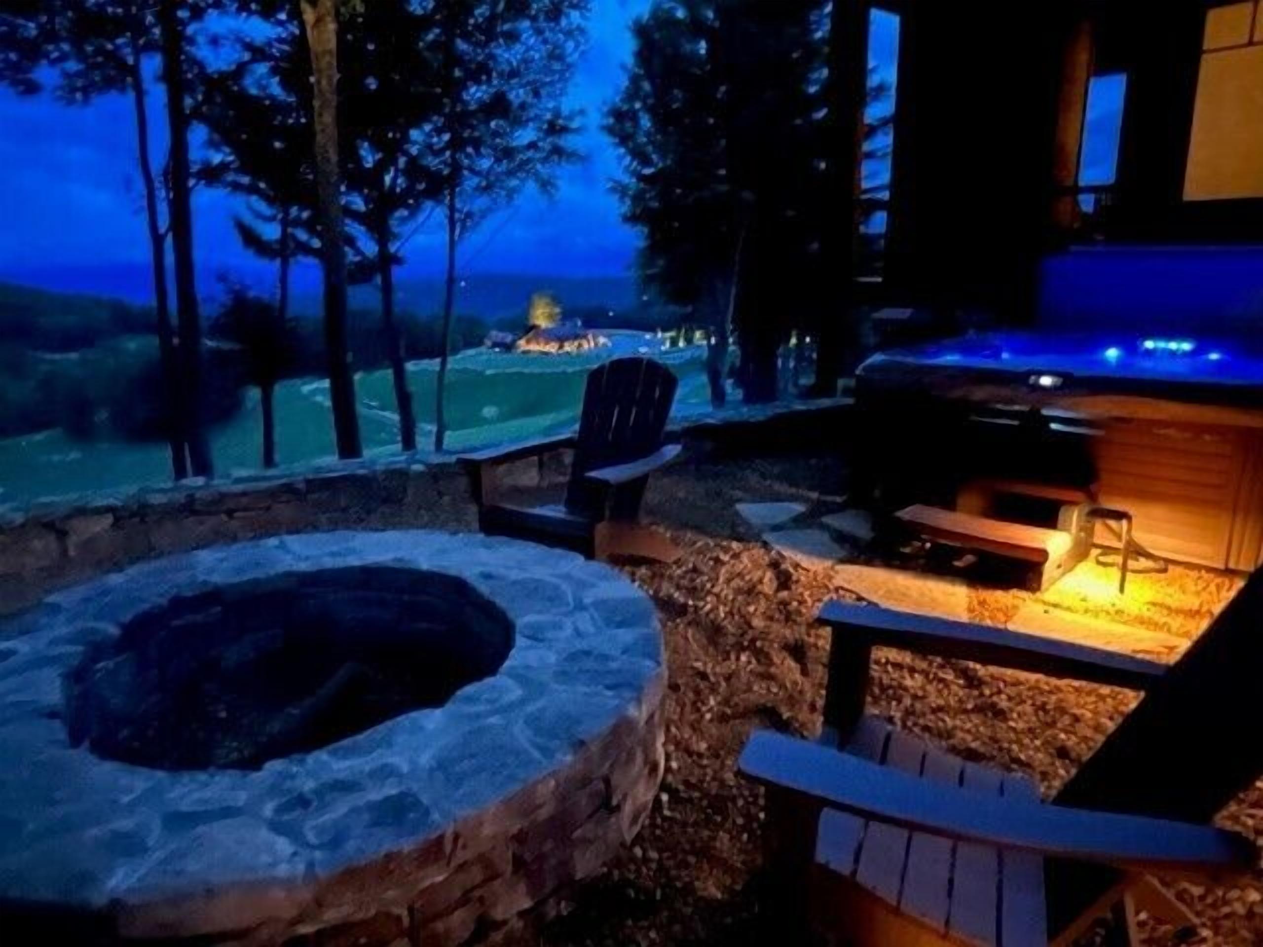 A stone fire pit with a chair, a grill area with a blue light, and a view of a forested area at dusk.