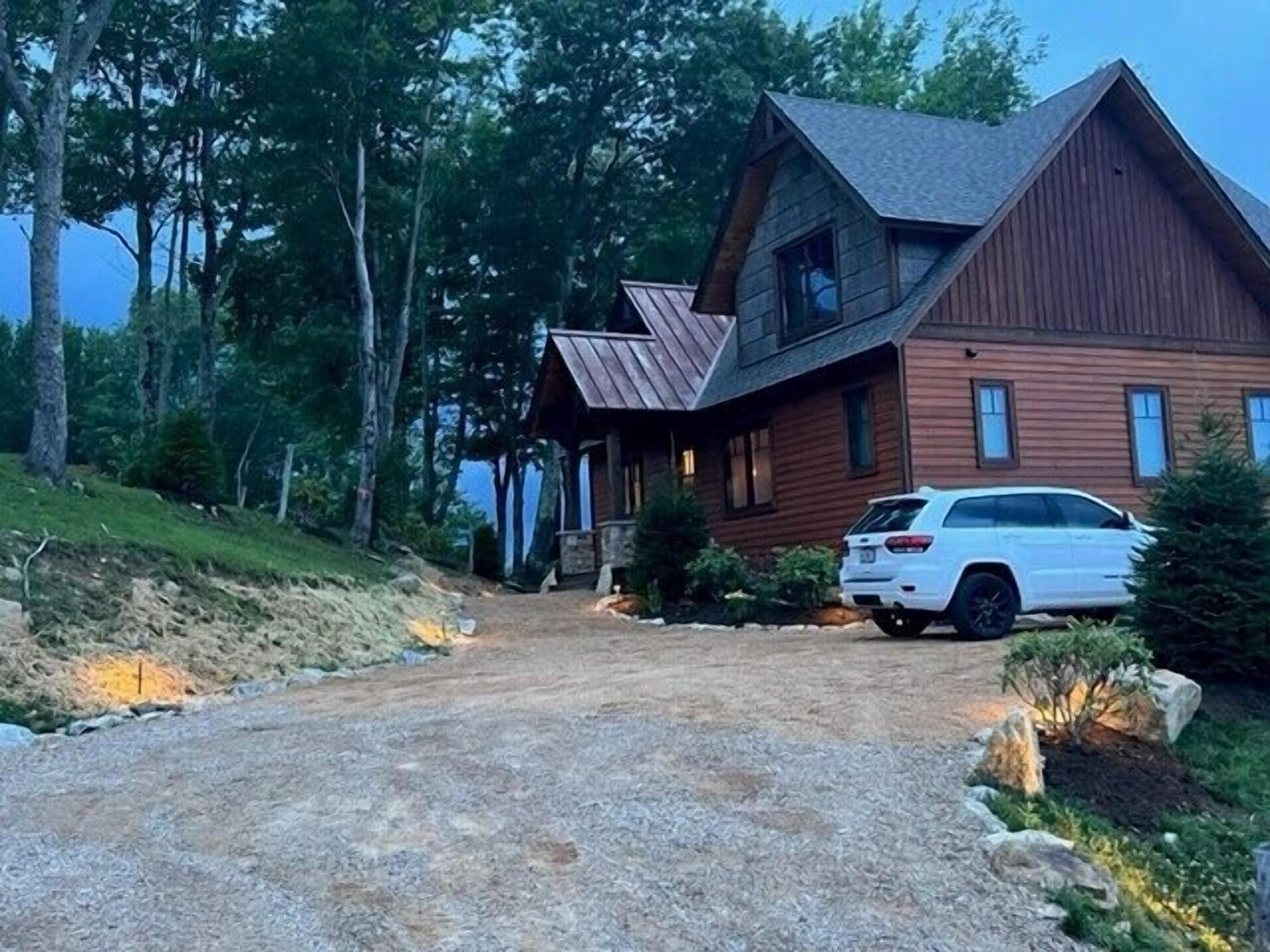 Una casa a due piani in legno con tetto spiovente, un vialetto sterrato e un SUV bianco parcheggiato davanti.