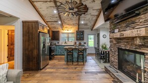 A rustic kitchen with a central island, wooden beams, a stone fireplace, and a large TV mounted on the wall. - Up the Creek (Linville)