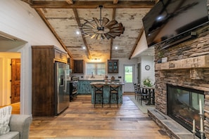 A rustic kitchen with a central island, wooden beams, a stone fireplace, and a large TV mounted on the wall. - Up the Creek (Linville)