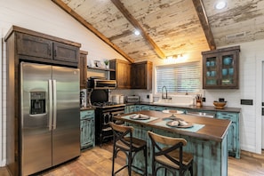 A kitchen with a stainless steel refrigerator, wooden cabinets, a central island with bar stools, and a rustic ceiling. - Up the Creek (Linville)