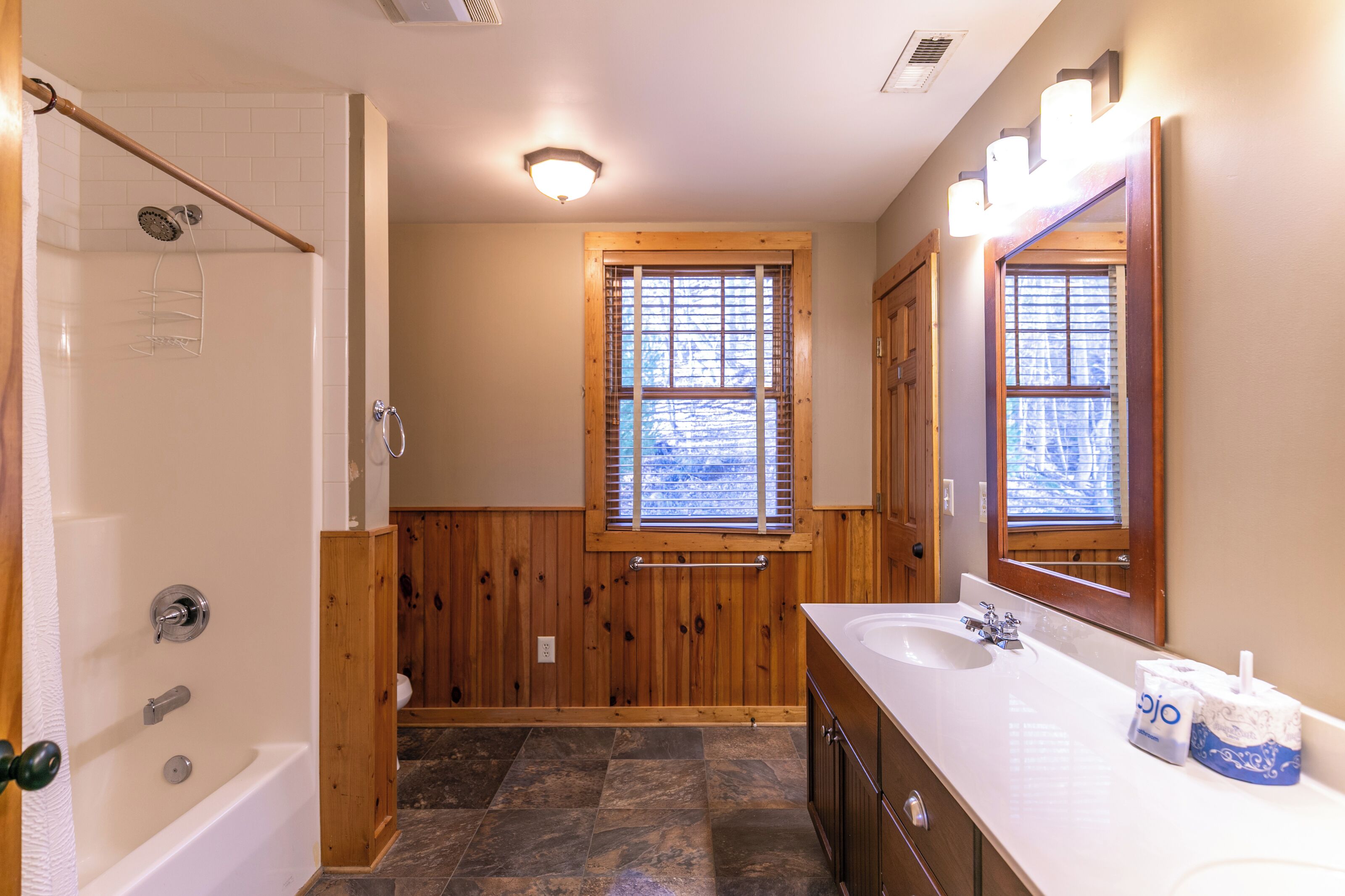 A bathroom with a bathtub, a wooden vanity with a mirror, and a window with blinds.