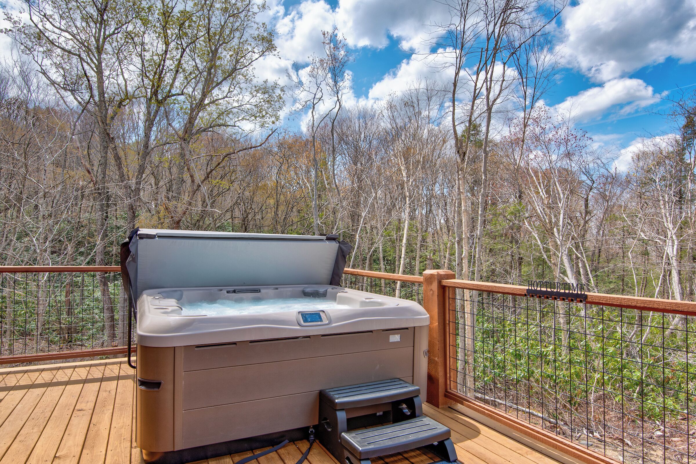 Un bain à remous sur une terrasse en bois, avec une rambarde en métal, entouré d’arbres.