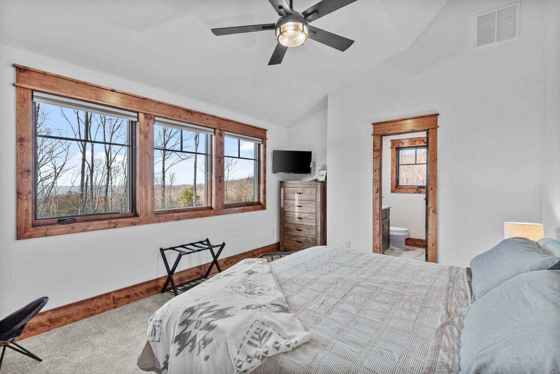 A bedroom with a large window, a ceiling fan, a dresser, a television, and a bed with patterned bedding.