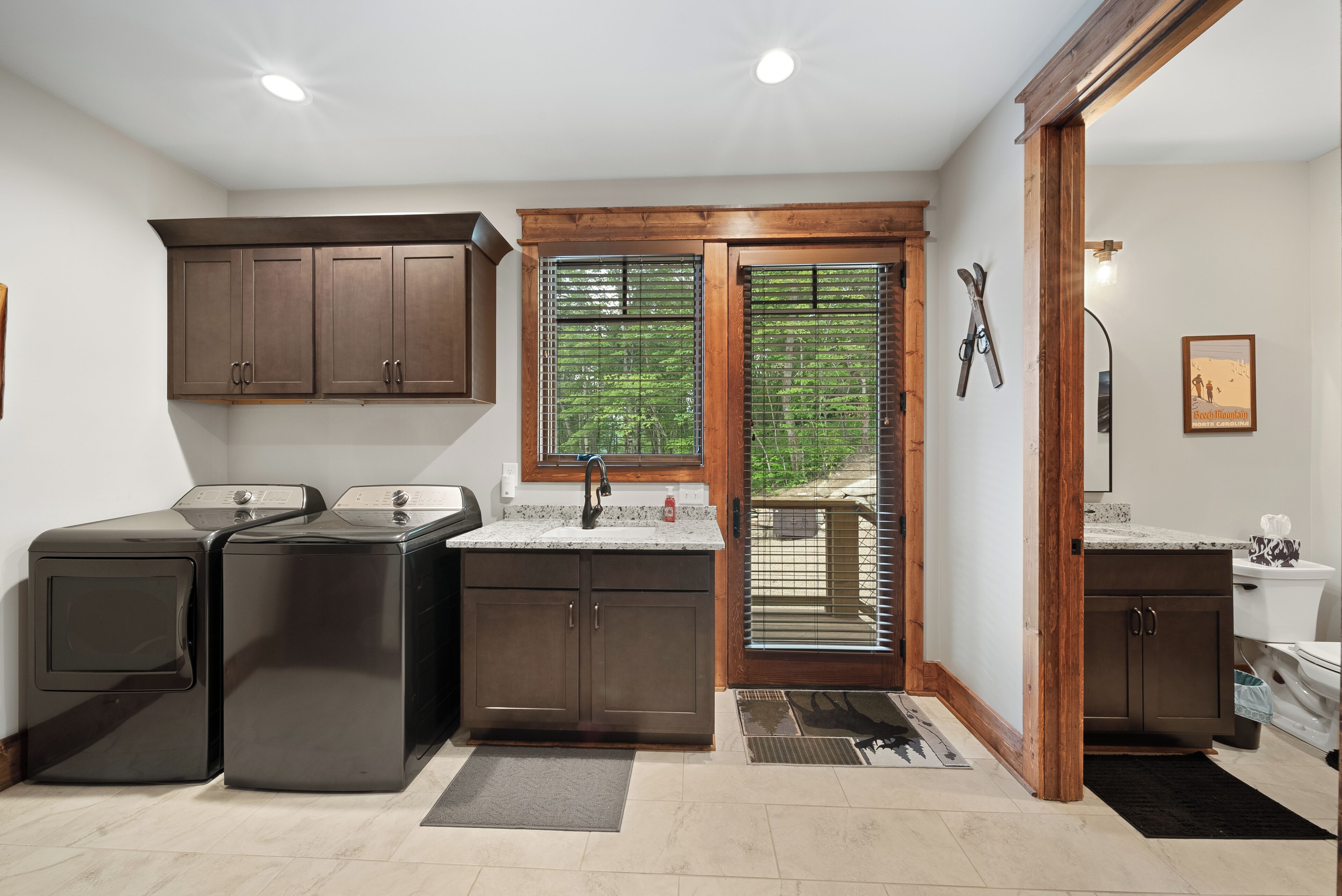 A laundry room with two front-loading washers and dryers, wooden cabinets, a window with blinds, and a door leading outside.