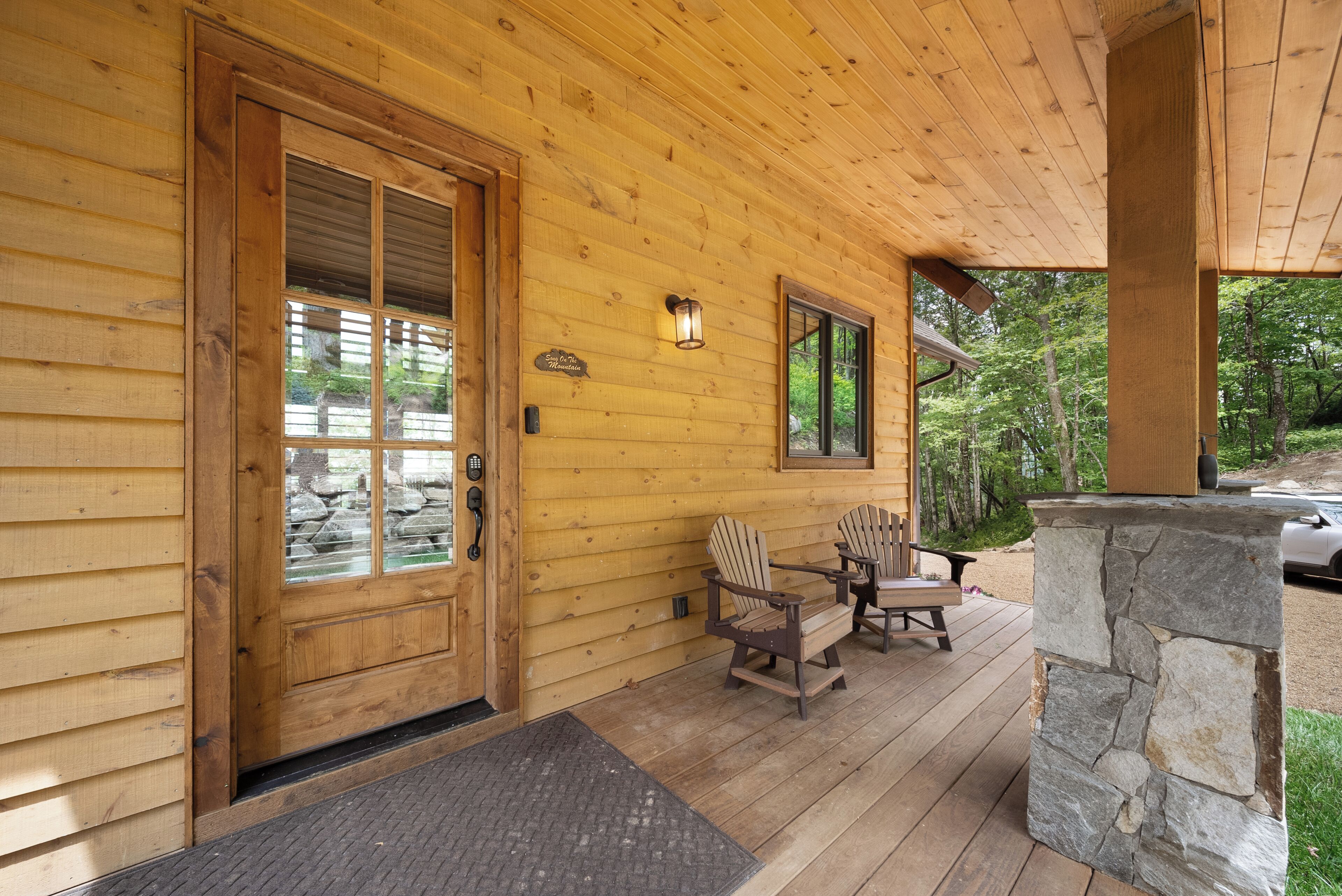A wooden cabin with a front door, two Adirondack chairs, a stone fireplace, and a wooden deck.