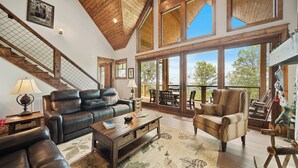 A spacious living area with a high ceiling, wooden beams, leather furniture, and large windows offering a view of the outdoors. - Song on the Mountain at Eagles Nest (Banner Elk)