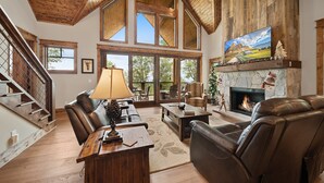 A spacious living room with a fireplace, leather sofas, a wooden coffee table, and a large window overlooking a scenic landscape. - Song on the Mountain at Eagles Nest (Banner Elk)