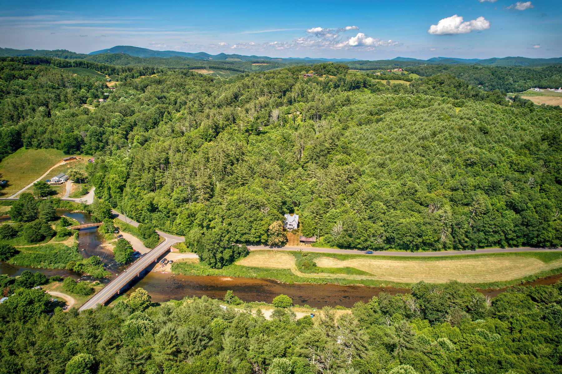 An aerial view of a forested area with a house surrounded by dense trees, a small bridge over a river, and a clear sky.