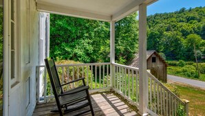 Eine Veranda mit Schaukelstuhl, weißen Geländern und Blick auf eine bewaldete Landschaft mit einem kleinen Holzgebäude.