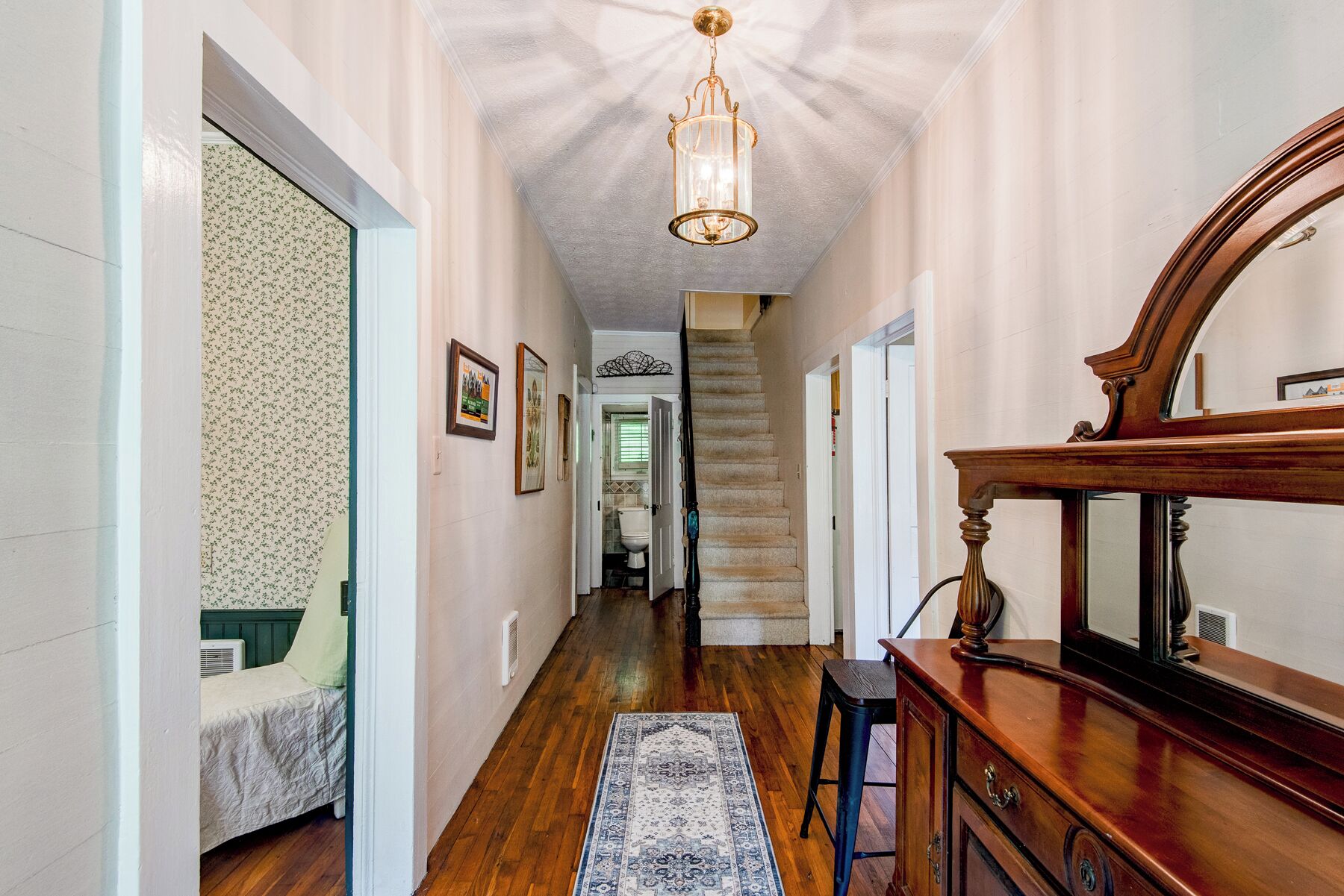 A hallway with a wooden floor, a rug, a staircase, and a mirror on a dresser.