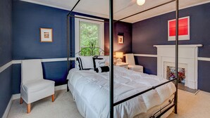 Ein Bett mit Baldachin, weißes Tuch, ein weißer Sessel, ein kleiner Tisch mit Lampe und ein Kamin.