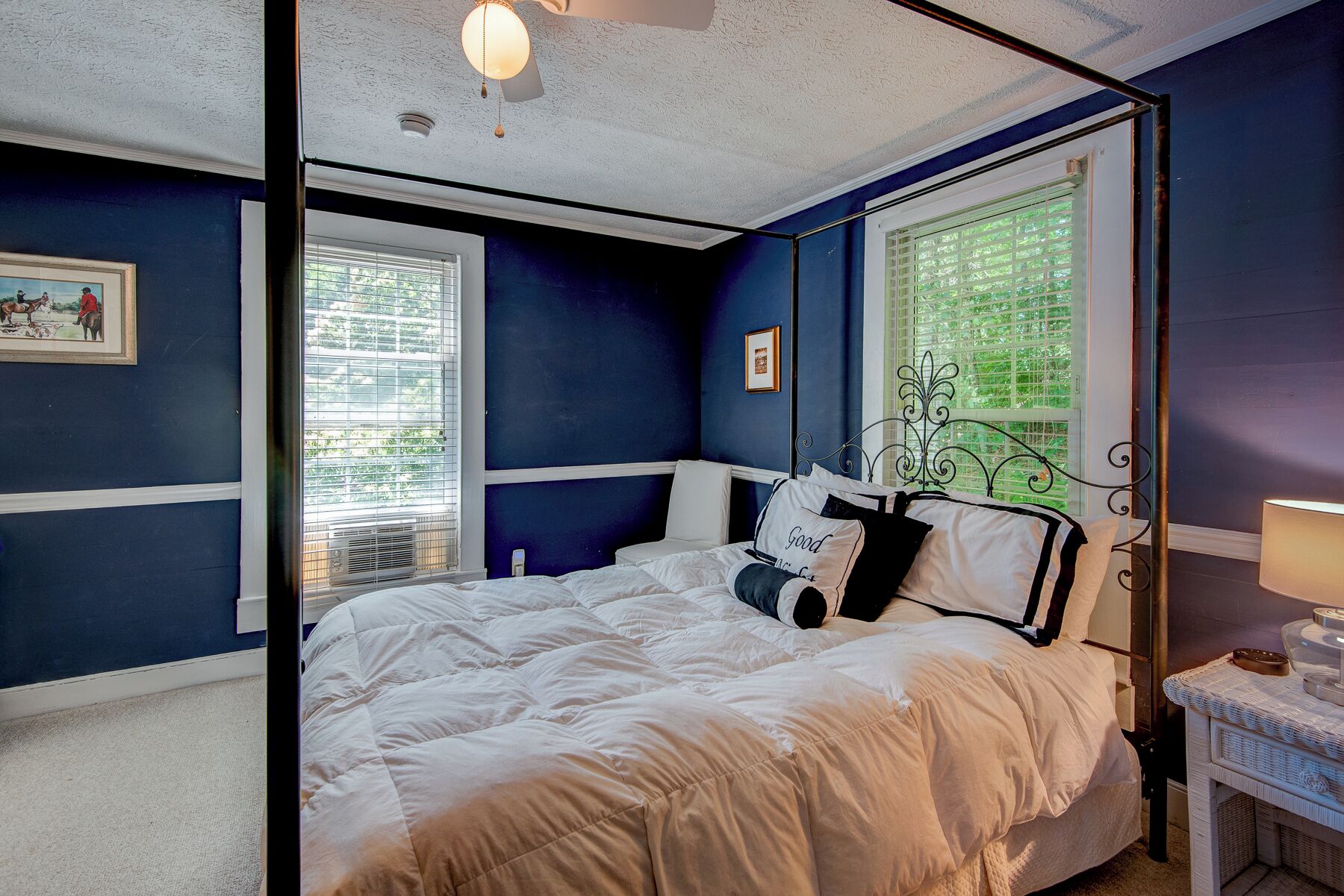 A bedroom with a bed, a window with blinds, a bedside table, and a wall-mounted lamp.
