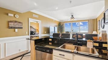 A modern kitchen with a black granite countertop, stainless steel sink, and white cabinetry. There is a dining area with two chairs and a large window offering a view of the ocean.