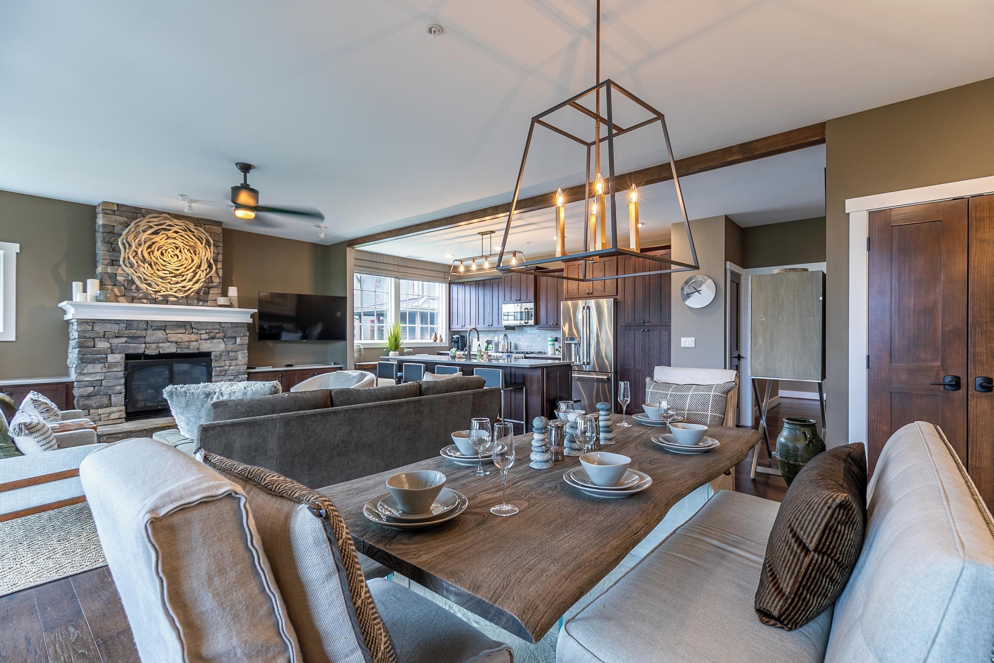 A modern living room with a fireplace, a dining area with a wooden table set for six, and a kitchen in the background.