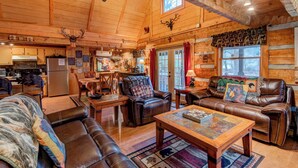 A cozy log cabin interior with a dining area, a fireplace, leather furniture, and a large window. - Bridgepoint Cabin Walk to Watauga River, hot Tub, Play in the Yard (Vilas)