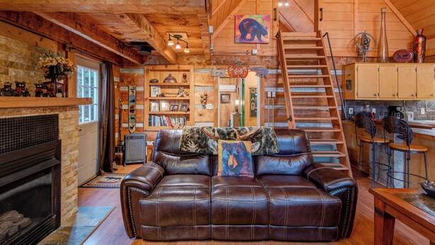 A cozy cabin interior with a fireplace, leather sofa, wooden shelves, and a staircase leading to an upper level. - Bridgepoint Cabin Walk to Watauga River, hot Tub, Play in the Yard (Vilas)