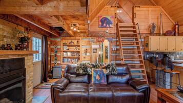 A cozy cabin interior with a fireplace, leather sofa, wooden shelves, and a staircase leading to an upper level.