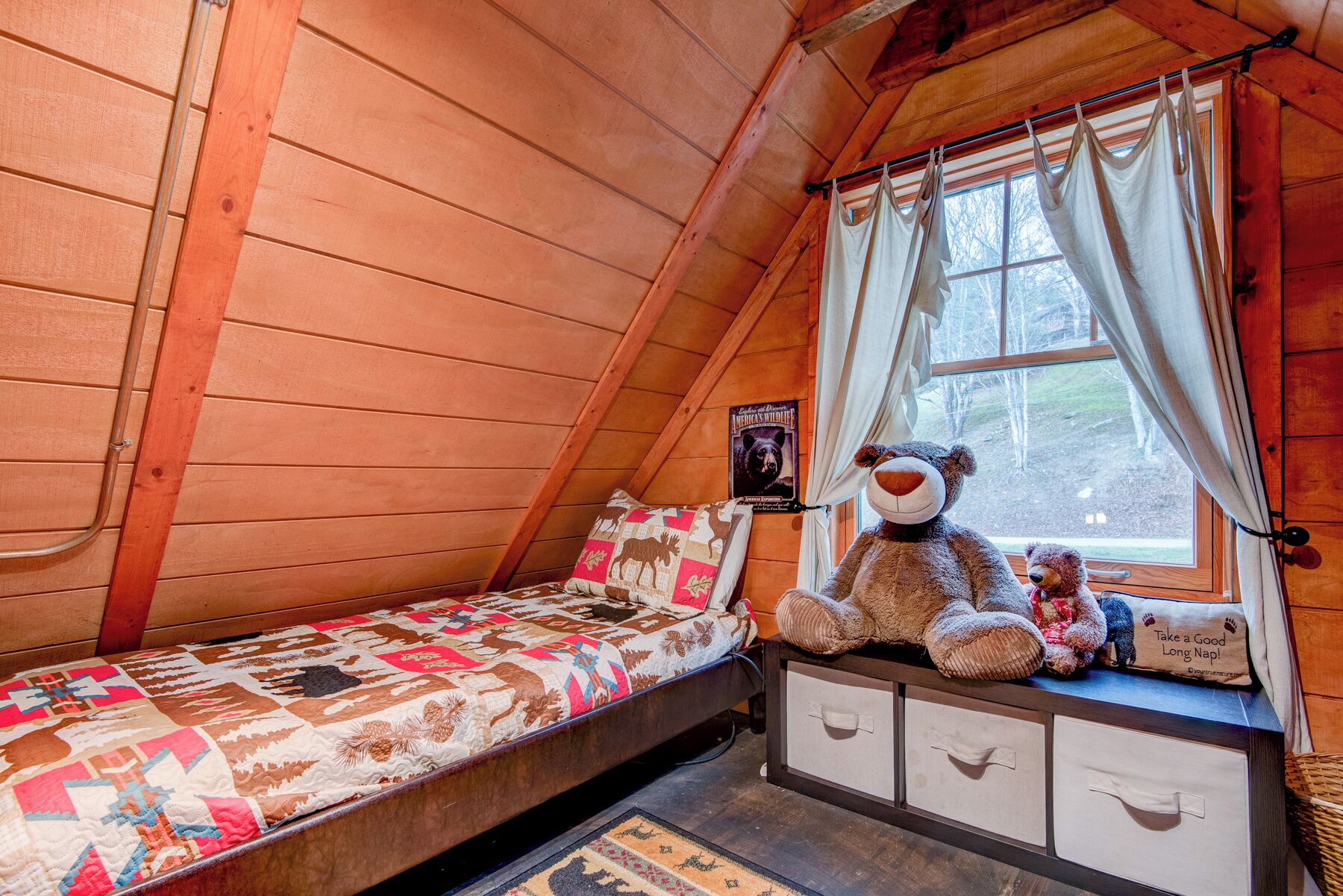 Een gezellig kamer in een blokhut met houten inrichting, een bed met sprei, een raam met gordijnen en een teddybeer op een plank.