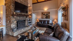 A cozy living room with a stone fireplace, leather sofa, and a television mounted on the wall. - Wolfman Lodge (Banner Elk)