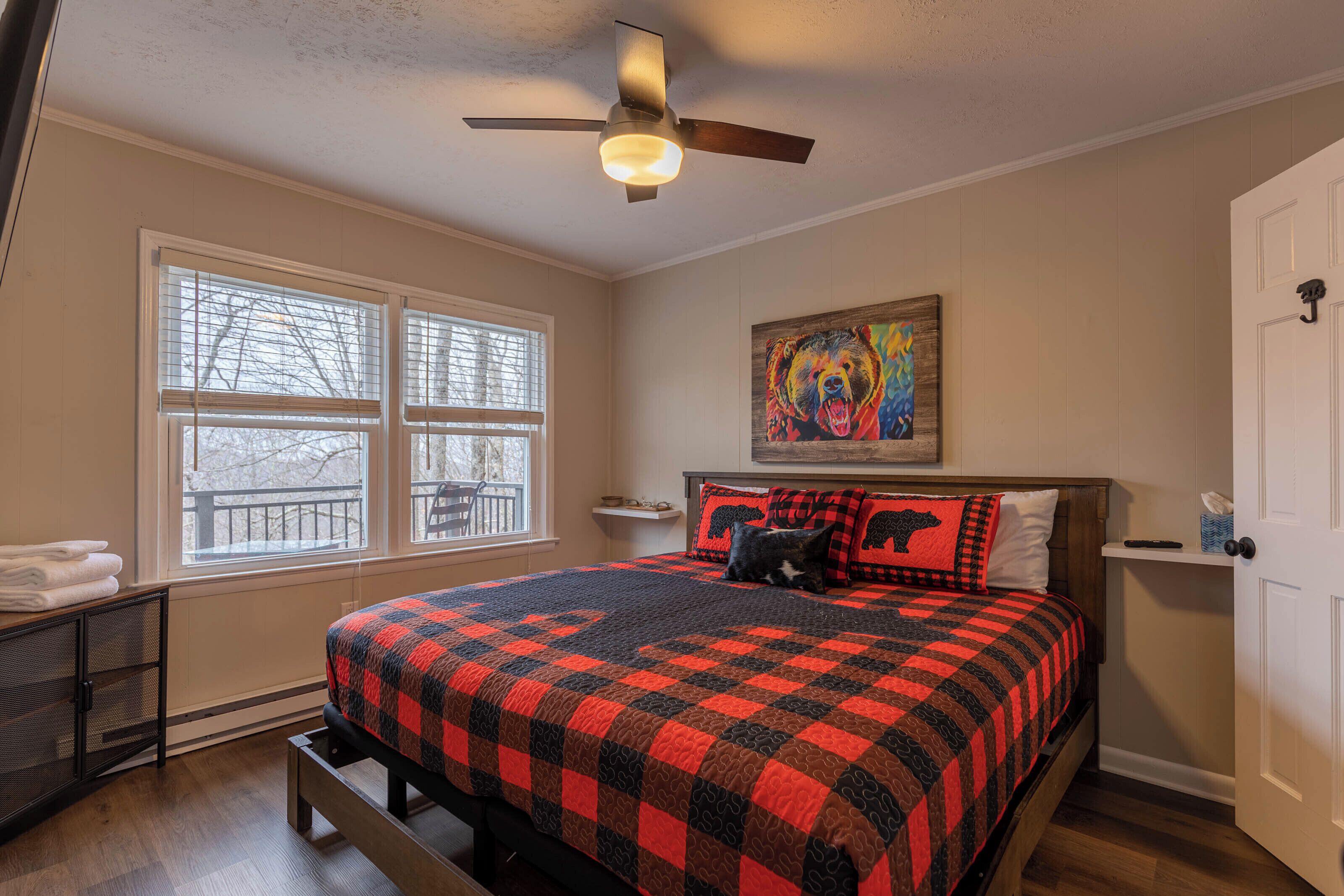 A bedroom with a bed, a ceiling fan, a window, a painting, and a door.