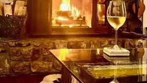 A stone fireplace with a lit fire, a glass of wine on a coaster, and a stone wall. - Wolfman Lodge (Banner Elk)