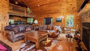 Ein geräumiger Innenraum einer Hütte mit Holzbalkendecke, einer großen Couch, einem Esstisch und einem Kamin.