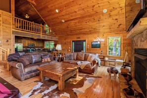 A spacious cabin interior with a wooden ceiling, a large couch, a dining table, and a fireplace. - Azalea Hill - Mountain Views, Hot Tub, Pool Table, Ping Pong (Boone)