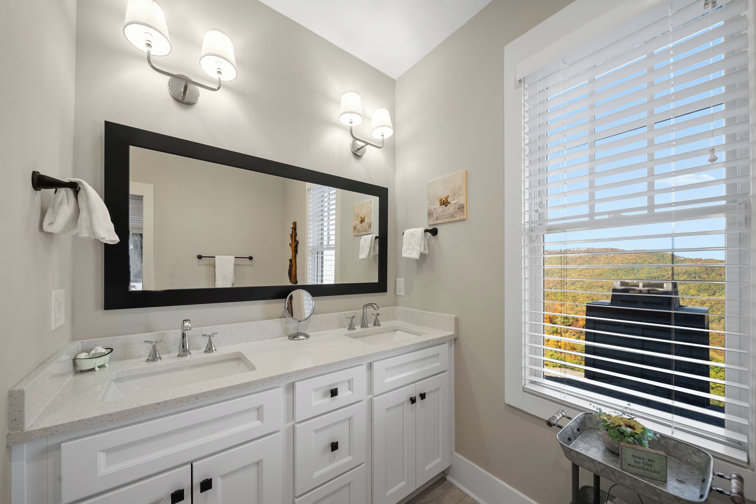 A bathroom with a double sink, a large mirror, wall sconces, a window with blinds, and a view of a mountain.